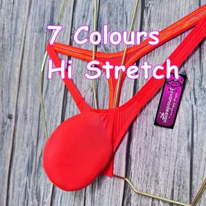 May include: A bright red thong swimsuit with a high-stretch design. The text "7 Colours Hi Stretch" is visible. A small tag with the brand name "Jovanadesign" is attached. The swimsuit is displayed on a gold-colored hanger against a wooden background.