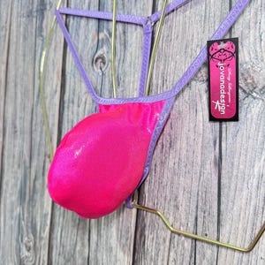 Könnte beinhalten: Ein leuchtend pinkfarbener String-Badeanzug-Unterteil mit lila Trägern. Das Kleidungsstück wird auf einem goldfarbenen Kleiderbügel vor einem Holzgrund gezeigt. Ein Etikett mit dem Text "jovanadesign" ist angebracht.