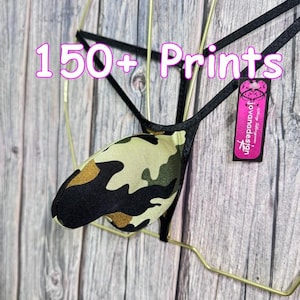 Op de afbeelding: Een camouflage-patroon string met zwarte bandjes, tentoongesteld op een goudkleurige hanger. De tekst "150+ Prints" is zichtbaar. Een roze label met de merknaam "jovanadesign" hangt aan het kledingstuk.