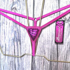 May include: A bright pink thong featuring thin elastic straps and a central triangular panel with a black and purple snakeskin pattern. A rectangular black tag attached to the side displays the text "Sexy Lingerie jovanadesign" in white.