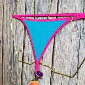 Può includere: Un perizoma bikini turchese con finiture e spalline rosa acceso. Il bikini presenta un ciondolo decorativo con una gemma viola e un ciondolo a forma di pesca. Il capo è esposto su una gruccia dorata su sfondo di legno.