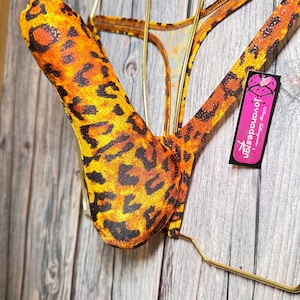 Puede incluir: Un traje de baño tanga con estampado de leopardo con un top a juego. El traje de baño es principalmente naranja y amarillo con manchas negras. Una etiqueta rosa con el texto "Jovandesign" está adjunta.