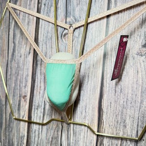 May include: A beige and mint green thong underwear displayed on a gold-coloured metal hanger. The underwear features a mint green mesh front panel and beige trim. A tag with the text "jovandesign" is attached.