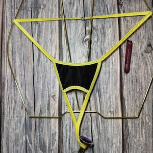 May include: A black and yellow thong-style lingerie with a decorative design. The lingerie is suspended from a gold-colored metal frame. A decorative plug with a purple top and a flame design is attached to the lingerie.