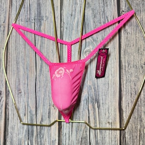 Puede incluir: Un tanga rosa brillante con un diseño floral en el panel frontal. El tanga está suspendido de una percha de metal dorado sobre un fondo de madera envejecida. Una pequeña etiqueta con el texto "jovanadesign" está adjunta.