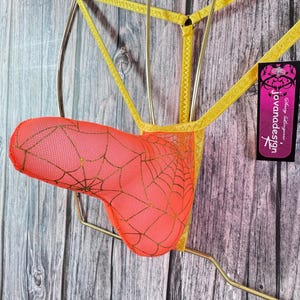May include: A neon orange pouch with a gold spiderweb pattern and yellow elastic straps. The garment features a black tag with the text 'jovanadesign' in white and pink lettering, attached to the side of the yellow waistband structure.