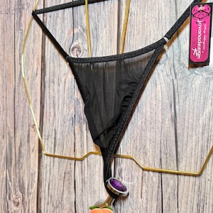May include: Black sheer thong with a decorative peach-shaped plug. The plug has a silver-colored metal base and a purple gem. The thong is hanging on a gold-colored metal display.