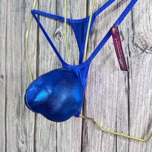 May include: A blue metallic thong swimsuit with a pouch. The swimsuit is displayed on a gold-colored hanger against a wooden background. A tag with the text "Jovanadesign" is attached.