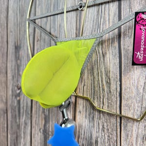 Può includere: Un tanga in rete verde lime con bordino argentato appeso a un appendiabiti in metallo dorato. Il tanga è etichettato "jovanadesign" e ha un oggetto a forma di stella blu appeso sotto.