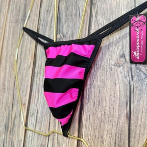 May include: A black and pink striped thong. The thong is displayed on a gold-colored hanger. A tag with the text "Jovana Design" is attached to the thong.