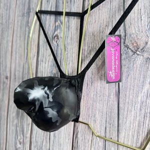 May include: Black camouflage-patterned lingerie with a g-string design. The lingerie is displayed on a gold-colored metal hanger against a wooden background. A tag with the text "jovanadesign" is attached.