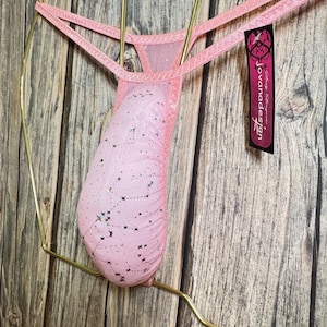 May include: Pink lingerie with a constellation pattern, hanging on a gold-colored metal hanger. The lingerie has a sheer fabric with a star design. A tag with the text "jovanadesign" is attached.