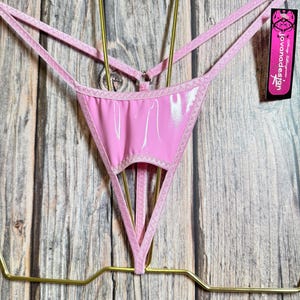 May include: Pale pink thong underwear with a glossy, wet-look appearance. The lingerie has delicate straps and a triangular front. A tag with the text "jovanadesign" is attached.