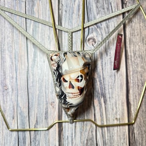 May include: A gray thong with a skull print. The skull design features black, white, and brown tones. The thong is displayed on a gold-colored metal hanger against a wooden background.
