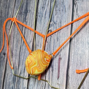 Puede incluir: Un tanga con estampado naranja y amarillo. El traje de baño tiene una pequeña bolsa con un patrón arremolinado de naranja y amarillo. Las correas son naranjas y el marco es dorado. El traje de baño se exhibe sobre una superficie de madera.