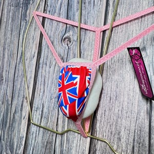 May include: Pink thong underwear featuring the Union Jack flag design, presented on a gold-coloured metal hanger. The underwear is displayed on a white form. A tag with "Jovana Design" is visible.