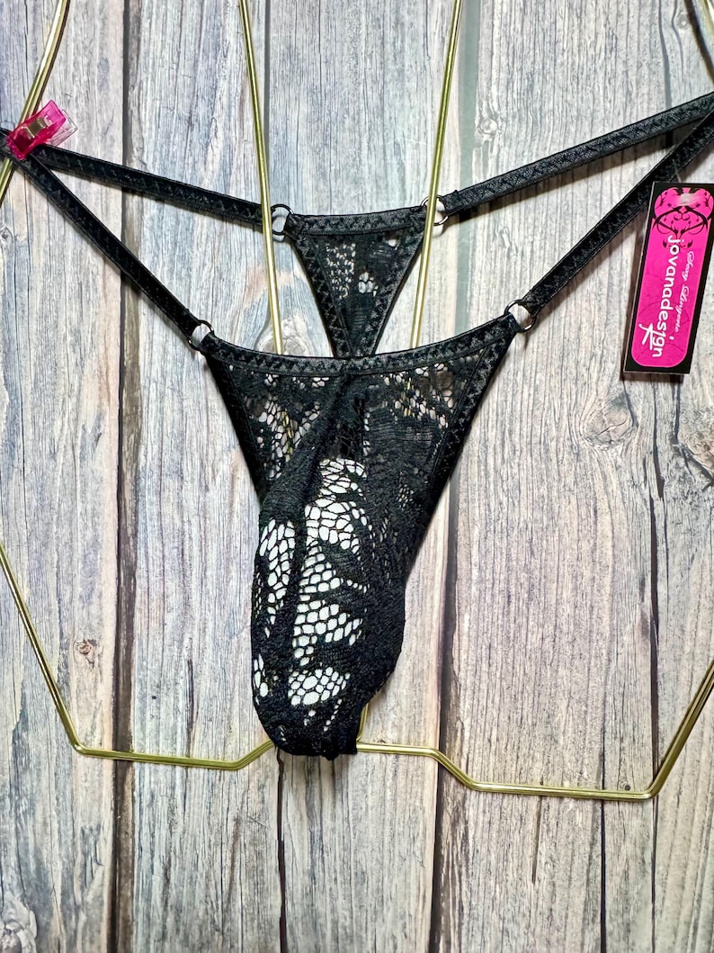 Puede incluir: Ropa interior de encaje negro con un patr&oacute;n floral. La ropa interior de estilo tanga tiene tirantes finos y detalles de anillos met&aacute;licos. Una etiqueta rosa con el texto "jovanadesign" est&aacute; adjunta.