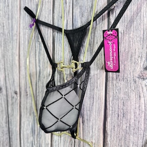 May include: Black lingerie with a sheer mesh pouch and diamond pattern. The lingerie has a gold-colored metal accent and a tag that reads "Javanadesign". The garment is suspended on a gold-colored wire hanger against a wooden background.