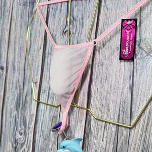 May include: A light pink thong with a white mesh pouch, hanging from a gold-colored geometric frame. A small, blue dolphin charm and a purple gem are attached to the thong. The tag reads "jovanadesign".