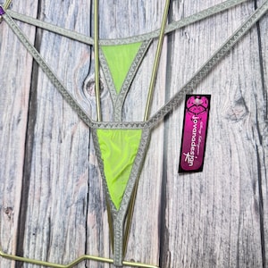 May include: A neon green and silver lingerie set. The set features a triangle-shaped design with a mesh fabric and silver trim. The lingerie is displayed on a gold-colored hanger. A tag with the text "Jovara Design" is visible.