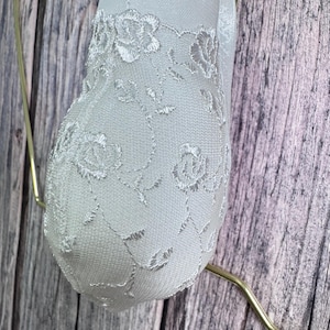 K&ouml;nnte beinhalten: Ein wei&szlig;es, herzf&ouml;rmiges Ornament mit zarten floralen Spitzendetails. Das Ornament wird von einem d&uuml;nnen, goldfarbenen Draht vor einem Hintergrund aus verwittertem, graut&ouml;nigem Holz gehalten.