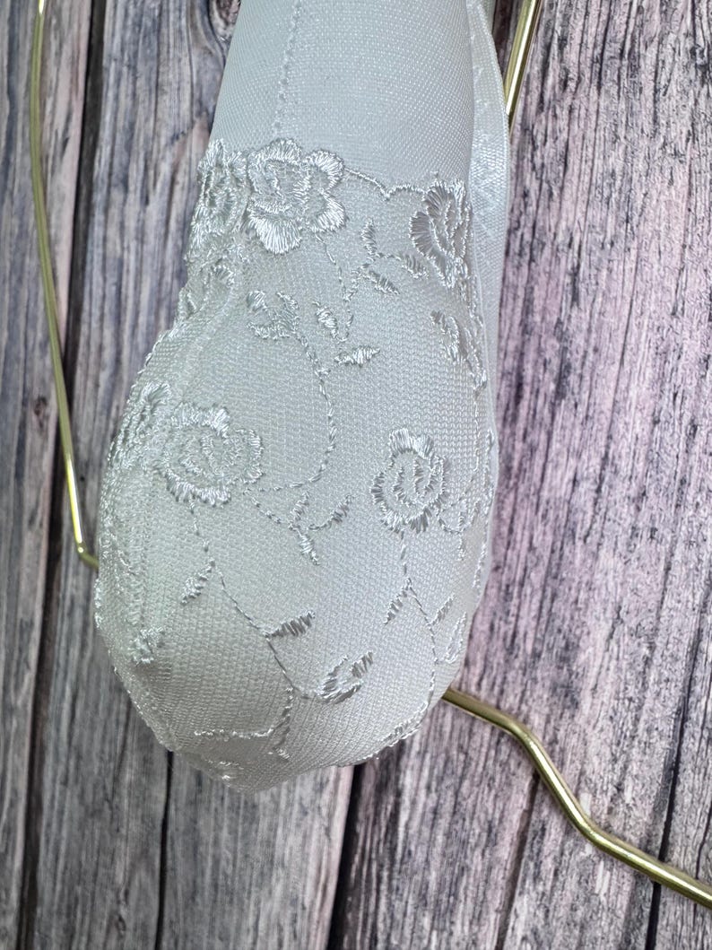 K&ouml;nnte beinhalten: Ein wei&szlig;es, herzf&ouml;rmiges Ornament mit zarten floralen Spitzendetails. Das Ornament wird von einem d&uuml;nnen, goldfarbenen Draht vor einem Hintergrund aus verwittertem, graut&ouml;nigem Holz gehalten.