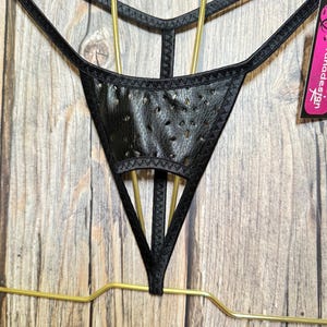 Puede incluir: Tanga negro con un panel frontal perforado similar al cuero y tirantes finos. La prenda está suspendida de una percha dorada sobre un fondo de madera. Una etiqueta con el texto "vanadesign" es visible.