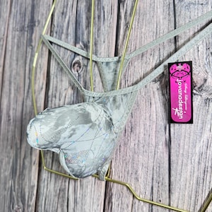 Puede incluir: Tanga plateado con una bolsa en forma de corazón, con un patrón brillante e iridiscente. La ropa interior se exhibe en una percha dorada sobre un fondo de madera. Una etiqueta rosa con el texto "jovanadesign" está adjunta.