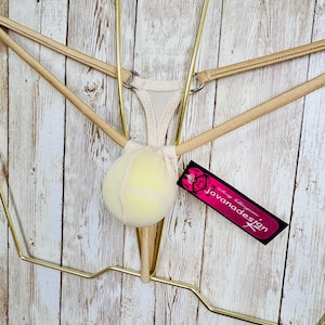 Op de afbeelding: Een beige string met een tennisbalvormig zakje in het midden, verbonden door dunne elastische bandjes en zilverkleurige metalen ringen. Een zwart rechthoekig label met roze tekst jovanadesign is aan de zijkant van het kledingstuk bevestigd.