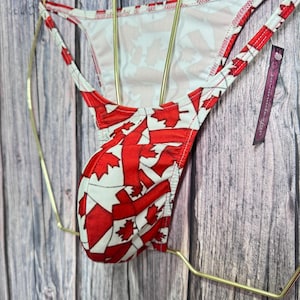 May include: Red and white patterned underwear featuring a maple leaf design. The underwear is displayed on a gold-colored hanger against a wooden background. The design is a repeating pattern of red maple leaves on a white background.
