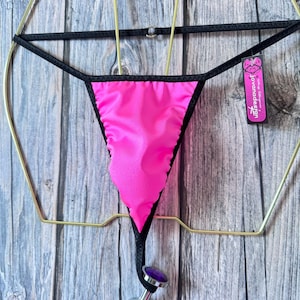 May include: A hot pink thong with black trim and a dolphin-shaped charm. The thong is suspended from a gold-colored metal hanger. A small tag with the text "lovandadesign" is attached.