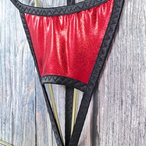 Könnte beinhalten: Roter, glitzernder String mit schwarzem Besatz und Trägern. Der String wird auf einem goldenen Kleiderbügel vor einem Holzgrund präsentiert. Der rote Stoff hat ein glänzendes, strukturiertes Aussehen.
