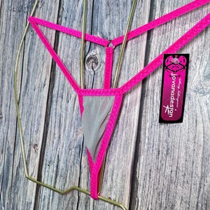Puede incluir: Un tanga rosa neón con un panel de malla gris. Los bordes están ribeteados con el mismo material rosa brillante. Se adjunta una etiqueta con el texto "Jovanadesign".