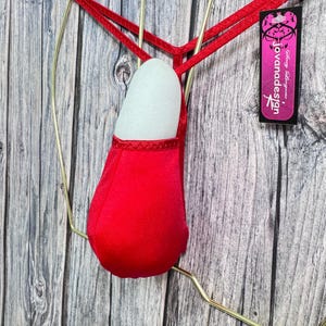 May include: A red thong-style lingerie piece with a pouch and thin straps. The lingerie is displayed on a gold-coloured hanger against a wooden background. A tag with the text "jovanadesign" is visible.