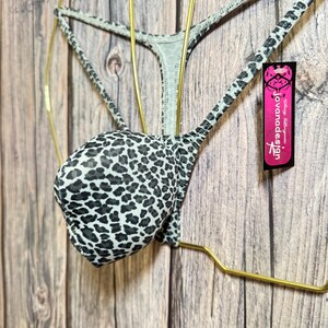 Op de afbeelding: Een zwart-wit lingerie set met luipaardprint, bestaande uit een bh en string, gepresenteerd op een goudkleurige hanger. De set heeft een herhalend dierenprintpatroon. Een roze label met "jovanadesign" is bevestigd.