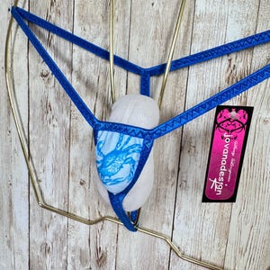 May include: A blue thong featuring a light blue front panel with a dark blue crab graphic. The garment has thin, textured blue straps and a black rectangular tag attached that reads "jovanadesign" in white text with a pink floral logo.