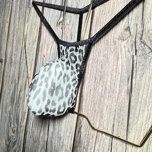 Puede incluir: Un tanga con estampado de leopardo en blanco y negro con ribete negro. El tanga se muestra en una percha dorada sobre un fondo de madera. La tela es transparente.