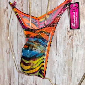 Op de afbeelding: Een string met een levendige, meerkleurige dierenprint met een glinsterende, gestippelde textuur. Het kledingstuk bevat een zwart label met roze accenten en de tekst "jovanadesign" die aan de zijkant hangt, weergegeven op een gouden draadkleerhanger.