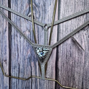 May include: A thong featuring a silver-toned elastic waistband and side straps. The front panel displays a metallic, iridescent scale-like pattern with black and silver hues. The garment is suspended on a gold-toned metal wire hanger.