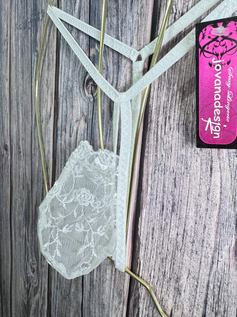 K&ouml;nnte beinhalten: Hellgr&uuml;ner String-Tanga mit floraler Spitze vorne. Der String h&auml;ngt an einem goldfarbenen Kleiderb&uuml;gel. Ein pinkfarbenes Etikett mit dem Text "Jovanadesign" ist sichtbar.