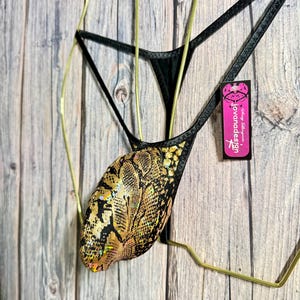 Op de afbeelding: Zwarte en gouden string-lingerie met slangenprint. De lingerie heeft een metallic gouden slangenpatroon op de voorkant en zwarte bandjes. Een roze label met de tekst "jovanadesign" is aan de band bevestigd.