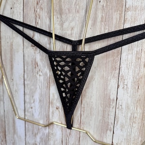 Puede incluir: Una tanga negra que presenta un panel frontal triangular con un patrón geométrico de rejilla abierta. La prenda está confeccionada con tiras elásticas finas y lisas que se conectan a la sección central recortada.