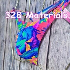 Puede incluir: Una braga de bikini con un vibrante estampado floral tropical en tonos rosa neón, azul brillante, morado y amarillo. La prenda incluye tiras laterales finas y muestra el texto 328 Materials impreso en rosa en la parte frontal.