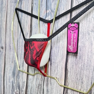 May include: Red and black patterned lingerie with black straps, displayed on a gold-colored wire frame. The lingerie has a unique design and is set against a wooden background. A tag with the text "jovanadesign" is visible.
