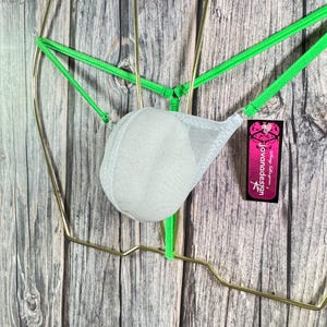 May include: A neon green and white lingerie set with a mesh pouch and thin straps. The lingerie is displayed on a gold-colored wire frame against a wooden background. A small tag with the text "jovanadesign" is attached.