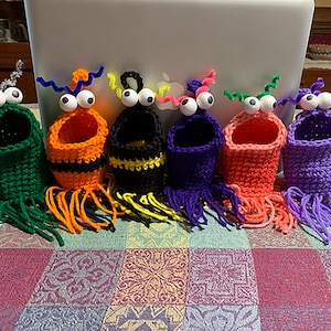 Może przedstawiać: Sześć dzianych ołówkowych pojemników w kształcie potworów z wypukłymi oczami i antenami z drucików. Pojemniki są w różnych kolorach, w tym zielonym, pomarańczowym, czarnym, fioletowym i różowym.