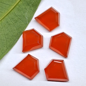 May include: Five faceted, shield-shaped carnelian gemstones with a vibrant orange-red hue. Each stone features a flat surface with geometric cut edges, arranged in a cluster on a white surface next to a single green leaf.