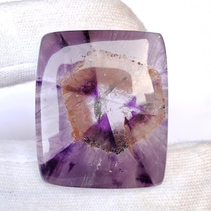 May include: A rectangular, polished amethyst gemstone with rounded corners. The stone features a translucent, light purple hue with a central, circular pattern of brown, white, and darker purple inclusions. The gemstone is held against a white background.