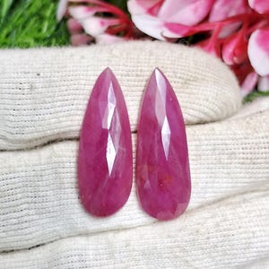 Op de afbeelding: Twee gefacetteerde, druppelvormige, dieproze edelstenen. De edelstenen hebben een gepolijst oppervlak met meerdere facetten die het licht reflecteren. De stenen worden gepresenteerd op een witte doek, met groene en roze bloemelementen op de achtergrond.