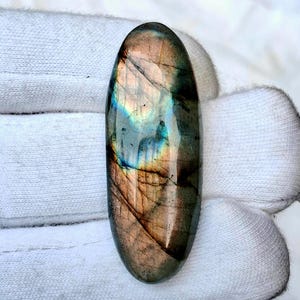 Può includere: Una pietra preziosa di labradorite di forma ovale con superficie lucida. La pietra mostra una miscela di colori, tra cui blu, verdi e marroni, con lampi iridescenti. La pietra misura circa 5 cm di lunghezza.